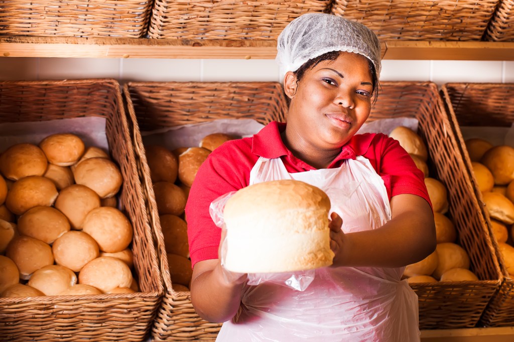 Mulher segurando pão nas mãos, rodeada de cestas de pães ao fundo