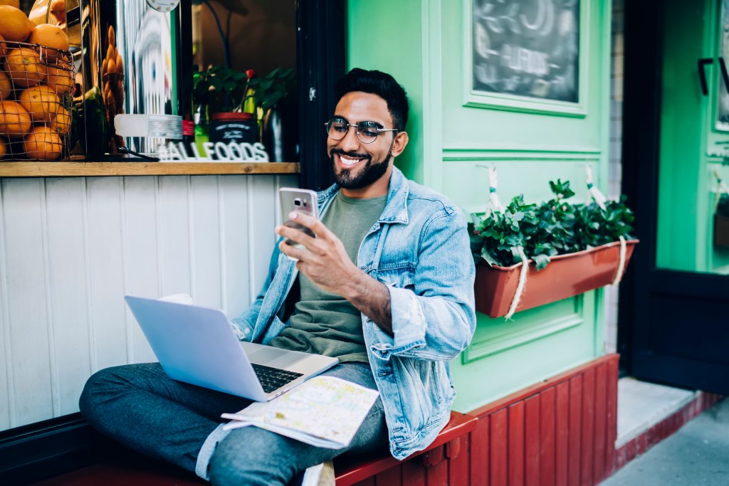 homem sorri sentando com computador no colo e celular na mão